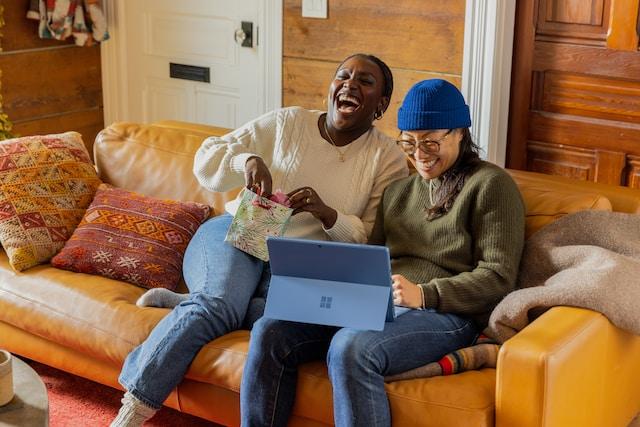 Image shows two people laughing. They're sat on a sofa with a device on their laps.